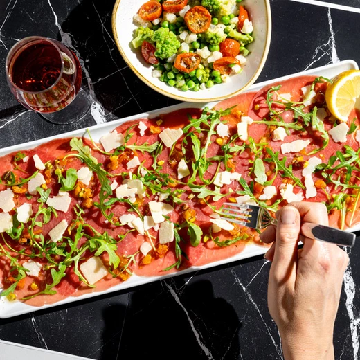 Fines tranches de bœuf* cru marinées, roquette, huile vierge à la tomate et à la pistache, copeaux de fromage italien. Suggestion de garniture : poêlée de légumes Fines tranches de bœuf* cru marinées, roquette, huile vierge à la tomate et à la pistache, copeaux de fromage italien. Suggestion de garniture : poêlée de légumes
